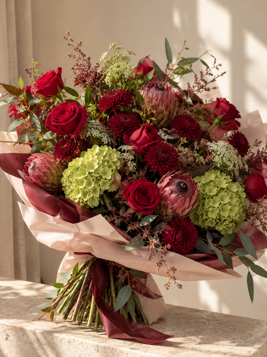 Ruby Garden hand-tied bouquet featuring red roses, burgundy chrysanthemums, pink proteas, green hydrangeas and eucalyptus in soft pink and burgundy wrapping.