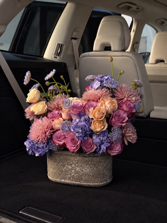 Pastel floral arrangement with pink and peach roses, lavender hydrangeas, pink chrysanthemums, and blue accent flowers in a textured ceramic vase.