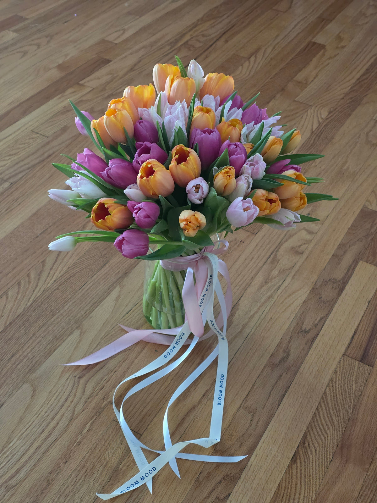 Tulip Crush arrangement of mixed pastel pink, lavender, peach, and apricot tulips in a clear glass vase with Bloommood signature ribbons.